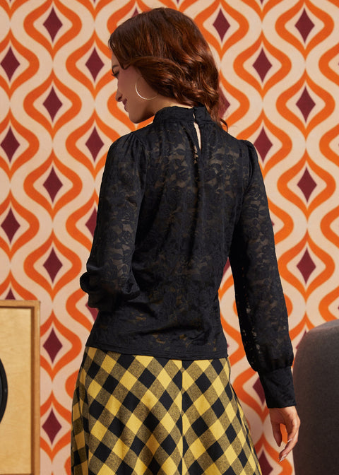 A woman shown from the back wearing a black lace long-sleeve blouse and a yellow and black checkered skirt, standing in front of a retro orange and cream patterned wallpaper.