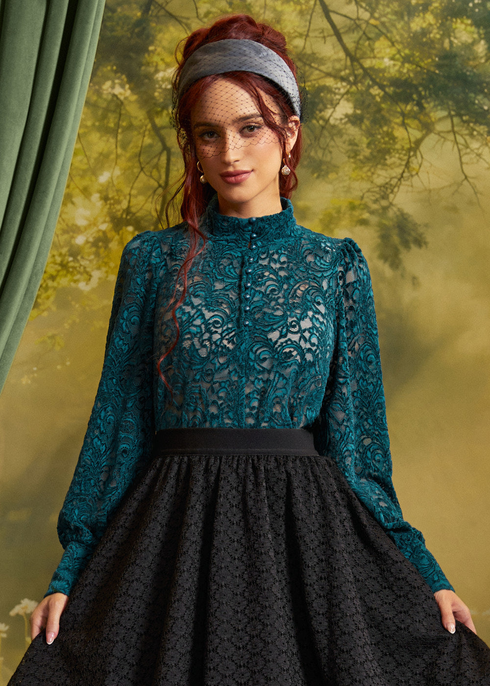 A woman modeling a long-sleeve lace blouse in teal with a black patterned skirt, styled with a gray headband and red hair, set against a vintage-style green and gold backdrop.