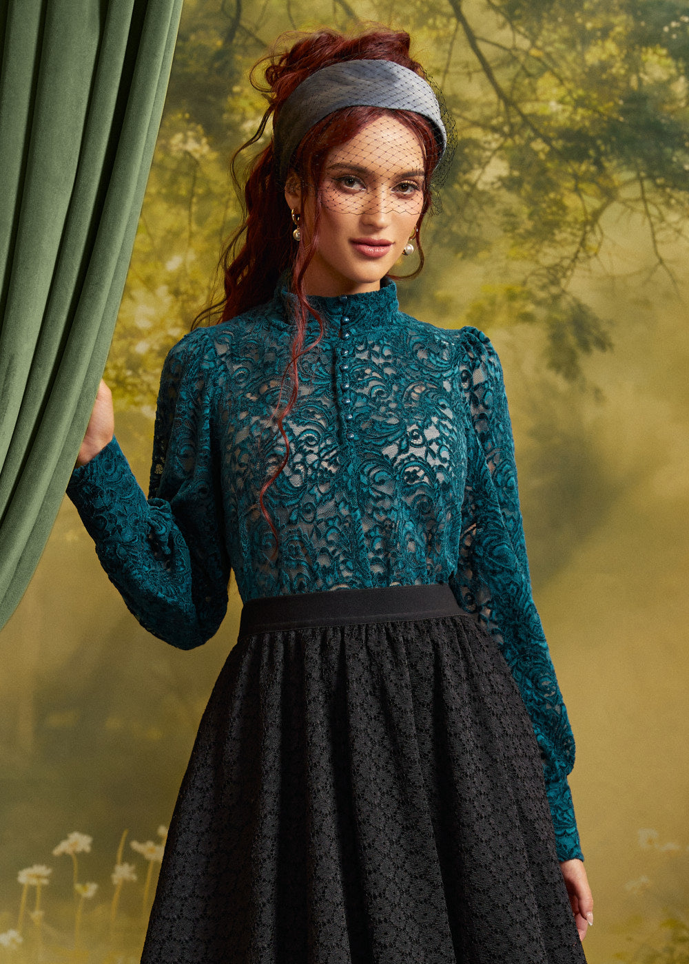 Woman wearing a teal floral lace blouse with high neckline and long sleeves, tucked into a black lace skirt, posed beside a green curtain.