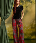 A woman modeling a black short-sleeve top with a dark collar and red plaid wide-leg pants, standing in front of a scenic forest backdrop with a green curtain on the left.