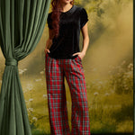 A woman modeling a black short-sleeve top with a dark collar and red plaid wide-leg pants, standing in front of a scenic forest backdrop with a green curtain on the left.
