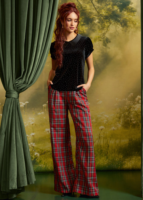 A woman modeling a black short-sleeve top with a dark collar and red plaid wide-leg pants, standing in front of a scenic forest backdrop with a green curtain on the left.