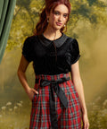 A woman modeling a black short-sleeve top with a Peter Pan collar and red plaid high-waisted trousers with a black waist tie, set against a soft-focus green and yellow forest backdrop.