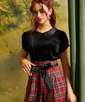 A woman modeling a black short-sleeve top with a Peter Pan collar and small silver polka dots, paired with a red plaid skirt featuring a black ribbon bow at the waist, posed against a soft-focus green and yellow backdrop.