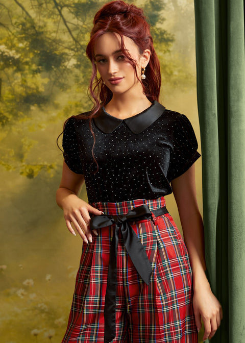 A woman modeling a black short-sleeve top with a Peter Pan collar and small silver polka dots, paired with a red plaid skirt featuring a black ribbon bow at the waist, posed against a soft-focus green and yellow backdrop.