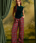 A woman stands in a studio setting wearing a dark green velvet sleeveless top and red plaid wide-leg trousers, posed in front of a scenic backdrop with green curtains.