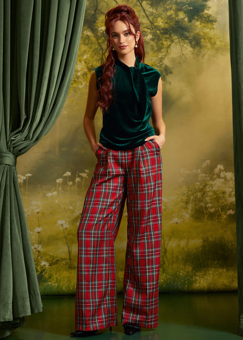 A woman stands in a studio setting wearing a dark green velvet sleeveless top and red plaid wide-leg trousers, posed in front of a scenic backdrop with green curtains.