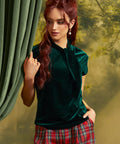 A woman with red hair models a dark green velvet short-sleeve top with a high neck and bow detail, paired with red and green plaid pants, standing next to a green curtain.