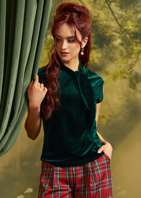 A woman with red hair models a dark green velvet short-sleeve top with a high neck and bow detail, paired with red and green plaid pants, standing next to a green curtain.