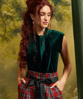 Model wearing a ModCloth emerald green velvet tie-neck top tucked into high-waisted red plaid trousers with a black sash belt, showing the dramatic neckline and cap sleeves.