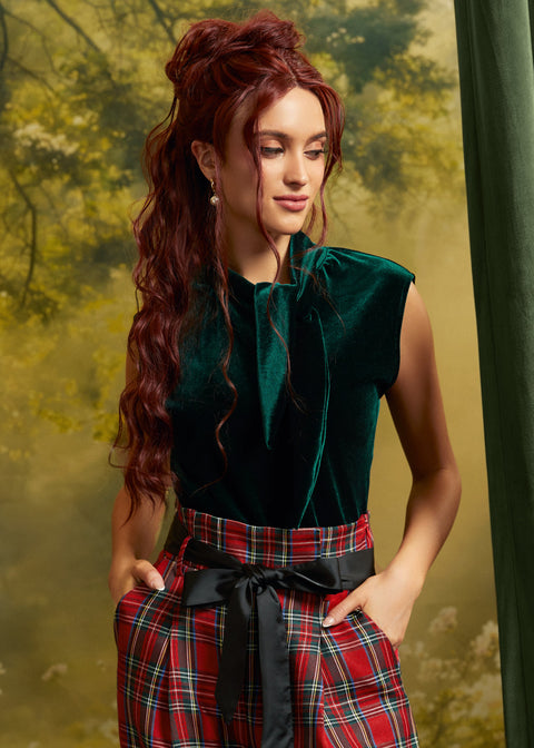 Model wearing a ModCloth emerald green velvet tie-neck top tucked into high-waisted red plaid trousers with a black sash belt, showing the dramatic neckline and cap sleeves.