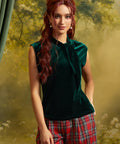 Model wearing an emerald green velvet sleeveless top with a dramatic tie neckline, styled with red plaid trousers against a garden-inspired backdrop.