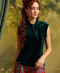 A woman with long wavy red hair wearing a dark green velvet sleeveless top with a high neck and bow detail, paired with a red and green plaid skirt, posed against a soft-focus forest backdrop.