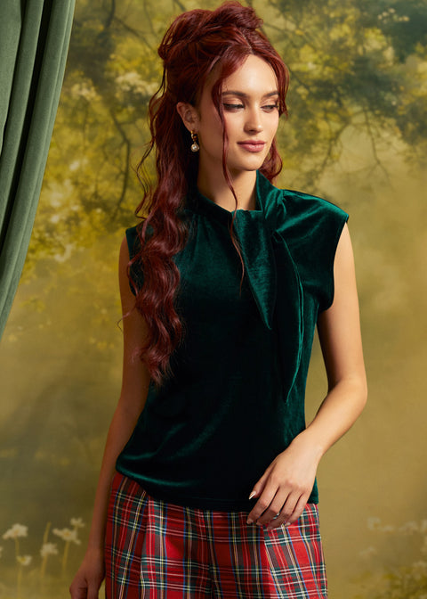 A woman with long wavy red hair wearing a dark green velvet sleeveless top with a high neck and bow detail, paired with a red and green plaid skirt, posed against a soft-focus forest backdrop.