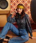 A woman sits on a carpeted floor in a retro-styled room, wearing a dark paisley long-sleeve blouse, blue wide-leg jeans, a mustard yellow headband, and black heels. A vintage speaker and a geometric orange-and-white wallpaper are visible in the background.