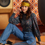 A woman sits on a carpeted floor in a retro-styled room, wearing a dark paisley long-sleeve blouse, blue wide-leg jeans, a mustard yellow headband, and black heels. A vintage speaker and a geometric orange-and-white wallpaper are visible in the background.