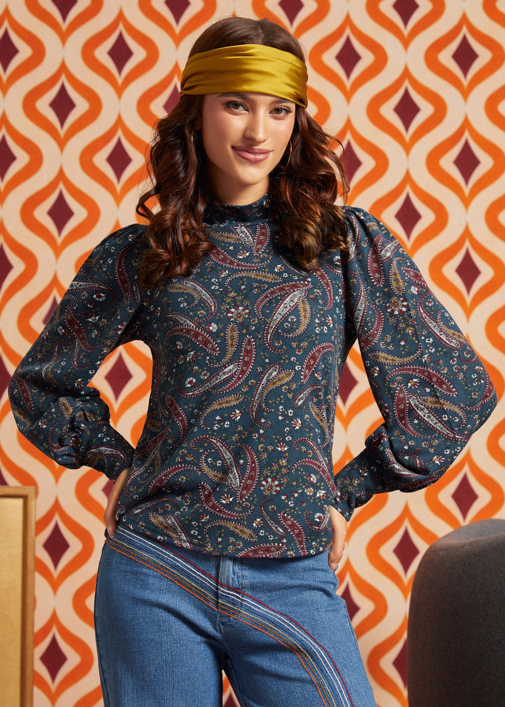A woman wearing a dark teal paisley long-sleeve top with blue jeans and a mustard yellow headband, posing in front of a retro orange and cream patterned wallpaper background.