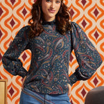 A woman wearing a dark teal paisley long-sleeve top with blue jeans and a mustard yellow headband, posing in front of a retro orange and cream patterned wallpaper background.