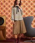 A woman models a patterned long-sleeve blouse with a black bow tie, paired with a brown midi skirt and burgundy lace-up ankle boots, set against a retro orange and white patterned wallpaper background.