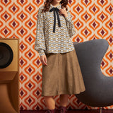 A woman models a patterned long-sleeve blouse with a black bow tie, paired with a brown midi skirt and burgundy lace-up ankle boots, set against a retro orange and white patterned wallpaper background.