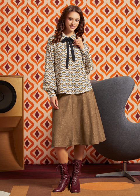 A woman models a patterned long-sleeve blouse with a black bow tie, paired with a brown midi skirt and burgundy lace-up ankle boots, set against a retro orange and white patterned wallpaper background.