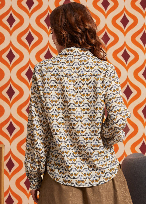 Woman wearing cream bird print blouse with brown skirt, back view showing long sleeves and shirttail hem