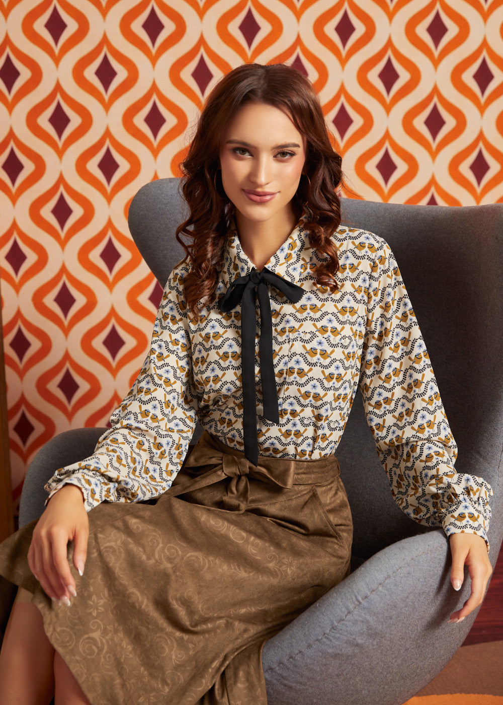 A woman sits in a gray armchair wearing a long-sleeve blouse with a brown, beige, and white bird pattern and a black bow at the collar, paired with a brown skirt. The background features retro orange and burgundy patterned wallpaper.