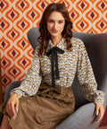 A woman sits in a gray armchair wearing a long-sleeve blouse with a brown, beige, and white bird pattern and a black bow at the collar, paired with a brown skirt. The background features retro orange and burgundy patterned wallpaper.