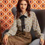 A woman sits in a gray armchair wearing a long-sleeve blouse with a brown, beige, and white bird pattern and a black bow at the collar, paired with a brown skirt. The background features retro orange and burgundy patterned wallpaper.