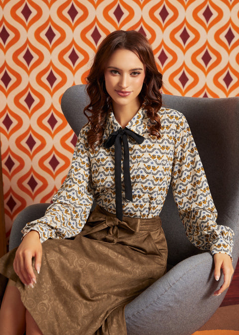 A woman sits in a gray armchair wearing a long-sleeve blouse with a brown, beige, and white bird pattern and a black bow at the collar, paired with a brown skirt. The background features retro orange and burgundy patterned wallpaper.