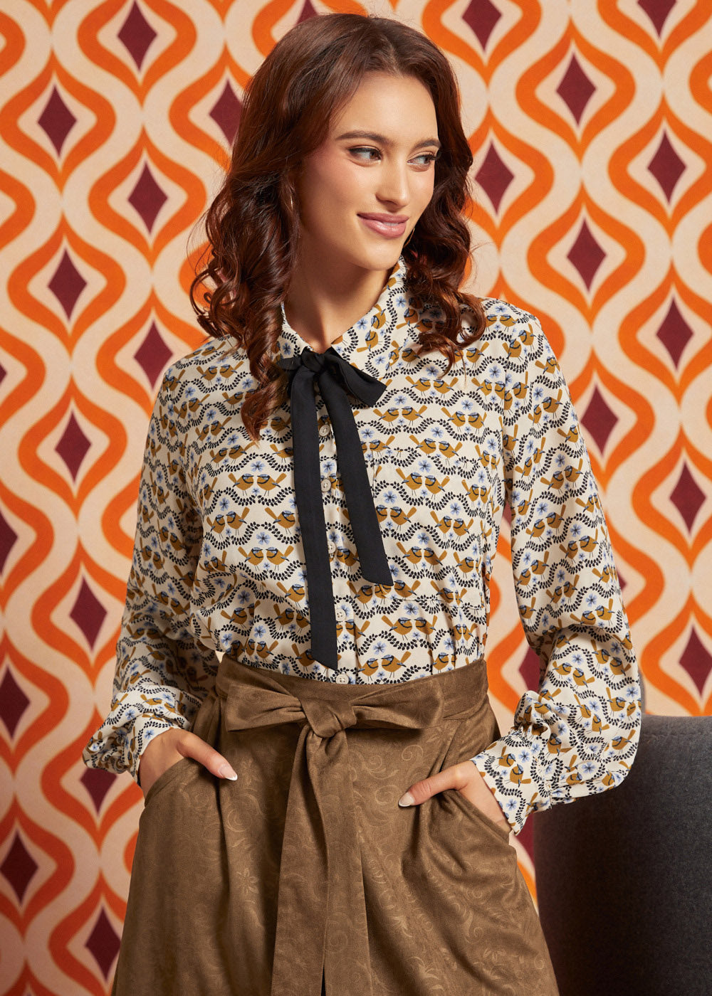 Woman wearing cream bird print collared blouse with black necktie and brown high-waisted skirt, front view with hands in pockets