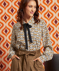 Woman wearing cream bird print collared blouse with black necktie and brown high-waisted skirt, front view with hands in pockets