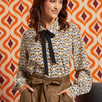 Woman wearing cream bird print collared blouse with black necktie and brown high-waisted skirt, front view with hands in pockets