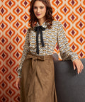 A woman models a long-sleeve blouse with a black bow tie-neck and a brown patterned skirt, posed against a retro orange and white patterned wall.