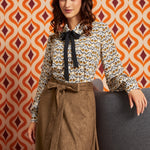 A woman models a long-sleeve blouse with a black bow tie-neck and a brown patterned skirt, posed against a retro orange and white patterned wall.