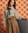 A woman models a long-sleeve blouse with a brown, beige, and white bird pattern and a black bow at the collar, paired with a brown patterned skirt, set against a retro orange and white patterned wall.