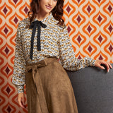 A woman models a long-sleeve blouse with a brown, beige, and white bird pattern and a black bow at the collar, paired with a brown patterned skirt, set against a retro orange and white patterned wall.
