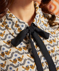 Close-up of cream bird print blouse showing pointed collar, black necktie, golden brown birds, blue flowers, and button front