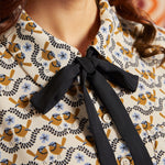 Close-up of cream bird print blouse showing pointed collar, black necktie, golden brown birds, blue flowers, and button front