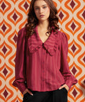 A woman wearing a long-sleeve, button-front blouse with a large ruffled collar, in a deep berry pink, paired with black trousers.
