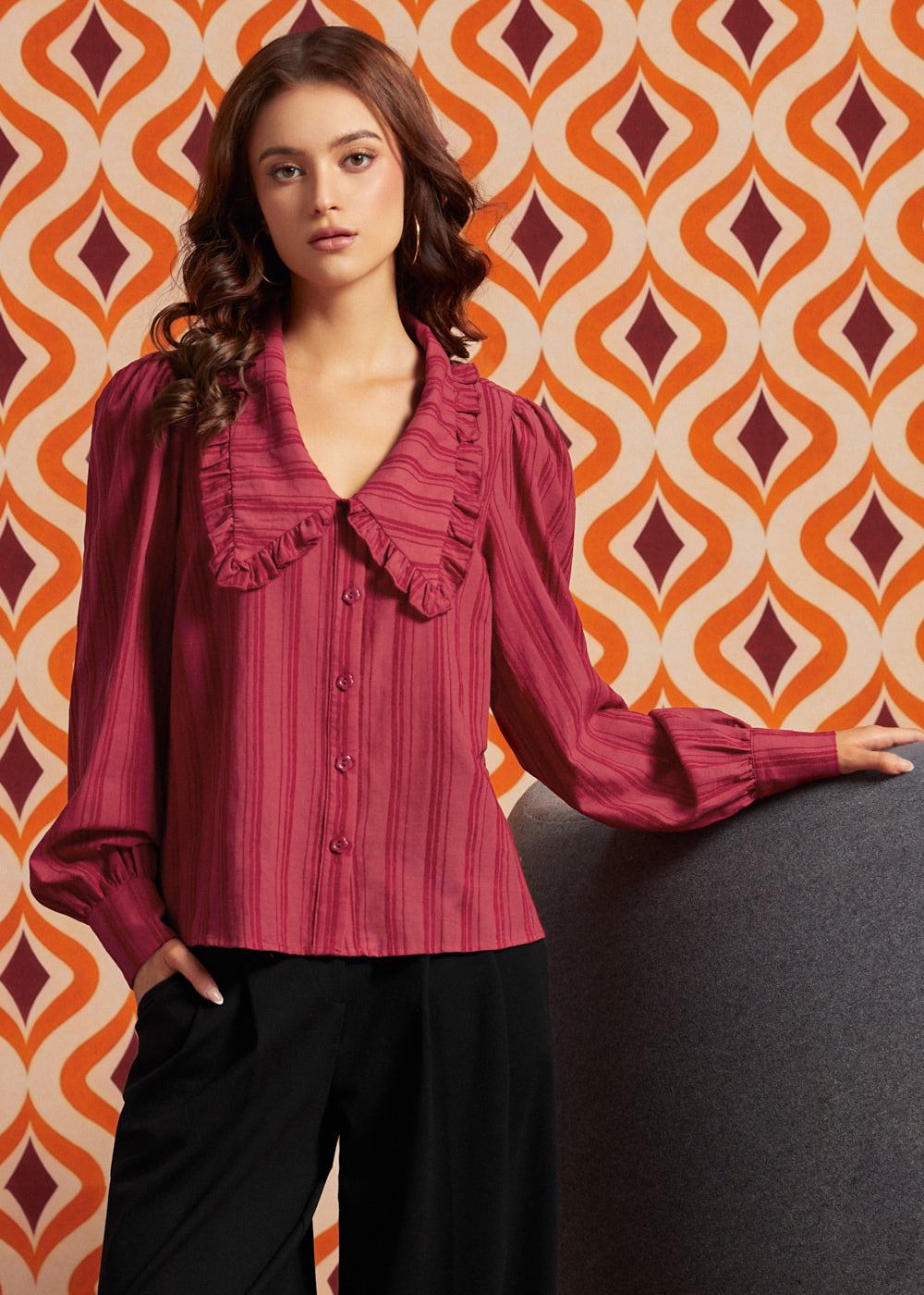 A woman wearing a long-sleeve ruffled blouse in a deep magenta pink with vertical stripes, paired with black pants