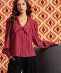 A woman wearing a long-sleeve ruffled blouse in a deep magenta pink with vertical stripes, paired with black pants