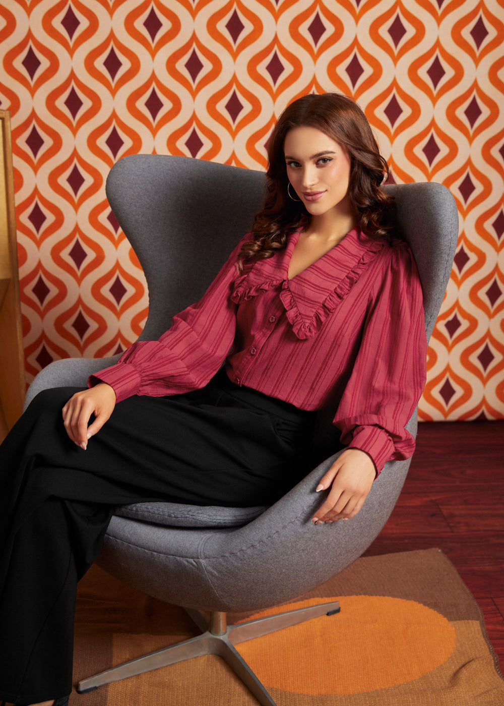A woman sits in a gray modern egg chair, wearing a dark pink ruffled blouse and black trousers from ModCloth