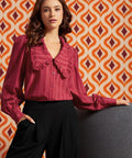 A woman models a long-sleeve, ruffled blouse in a deep berry pink with vertical stripes, paired with black wide-leg trousers, set against a retro orange and cream patterned wallpaper background.