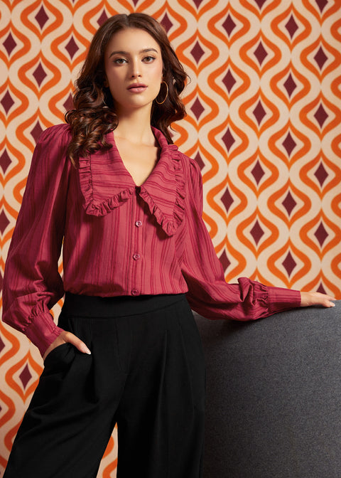 A woman models a long-sleeve, ruffled blouse in a deep berry pink with vertical stripes, paired with black wide-leg trousers, set against a retro orange and cream patterned wallpaper background.