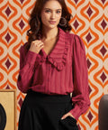 A woman wearing a long-sleeve, ruffled blouse in a deep magenta-pink color, paired with black trousers.
