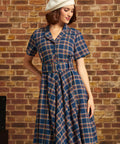 A woman wearing a short-sleeve plaid dress with a matching belt and a white beret, standing against a brick wall.