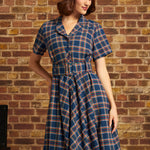 A woman wearing a short-sleeve plaid dress with a matching belt and a white beret, standing against a brick wall.