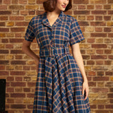 A woman wearing a short-sleeve plaid dress with a matching belt and a white beret, standing against a brick wall.