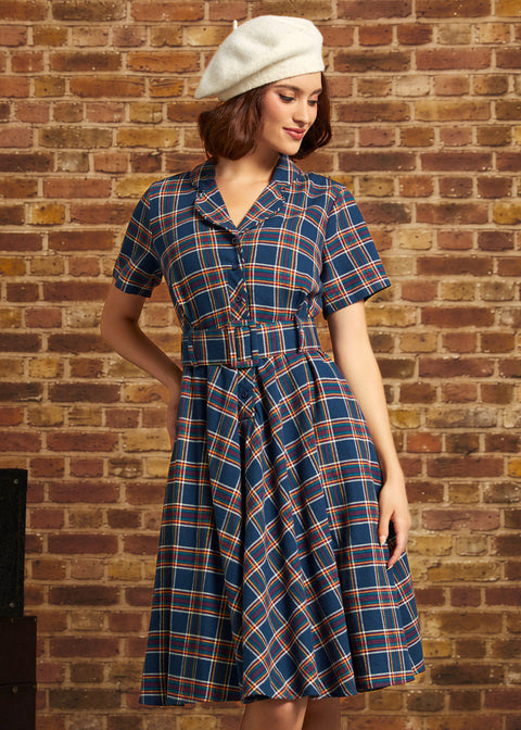 A woman wearing a short-sleeve plaid dress with a matching belt and a white beret, standing against a brick wall.